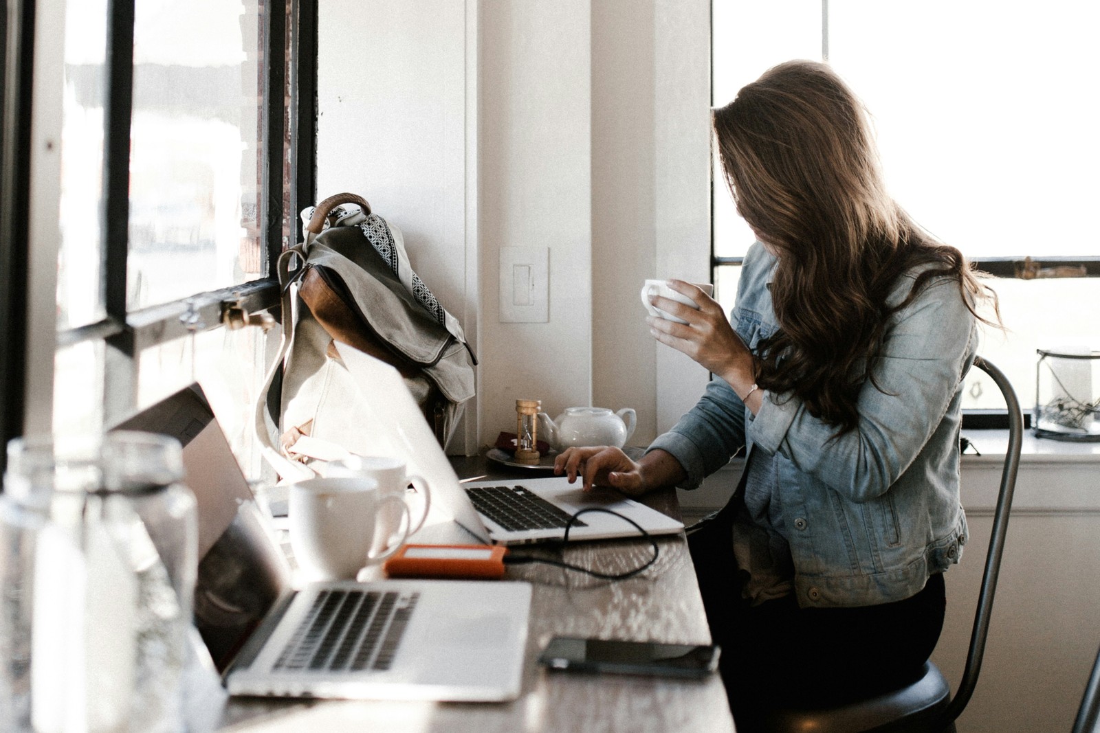 Woman at desk near a window, holding a cup, working on a laptop.