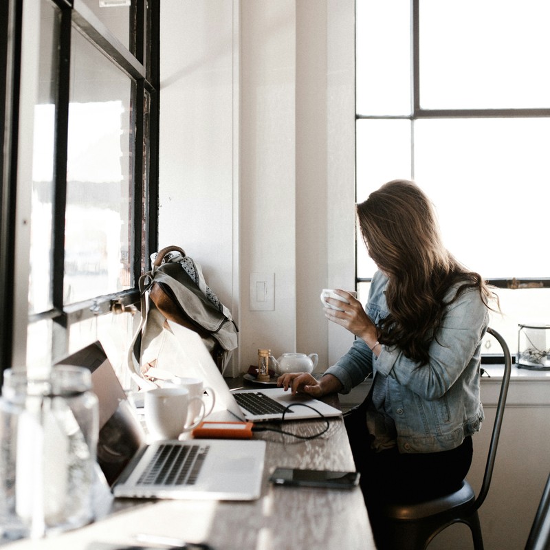 Woman at desk near a window, holding a cup, working on a laptop.